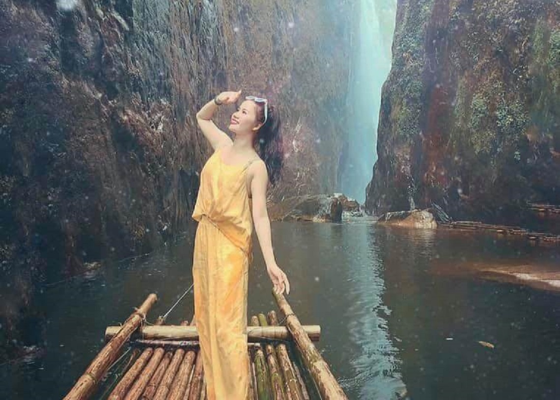 Bạn nữ mặc bộ jumset vàng đứng trên chiếc bè trôi dưới dòng nước giữa 2 khe vách núi đá